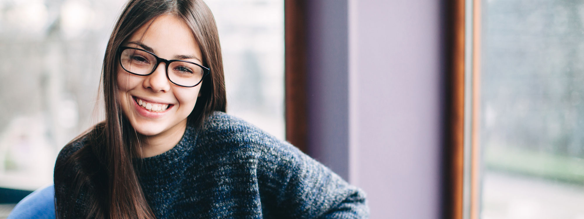 Teenage girl with glasses sitting in front of a window and smiling.